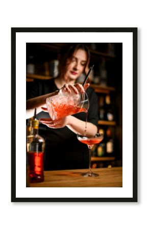 Cute young female bartender pouring cold brown cocktail from mixing glass into stemmed glass