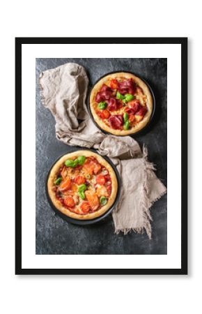 Two Traditional pizza with bresaola, smoked salmon, cheese, tomatoes and basil served on black plates with textile over black texture background. Top view with space.