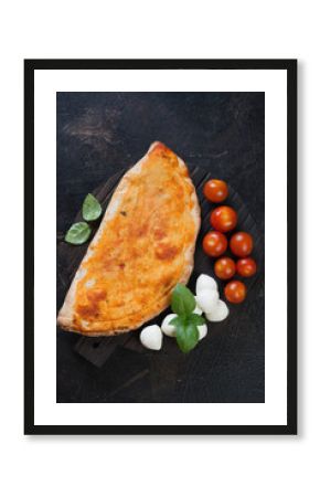 Above view of pizza calzone with cherry tomatoes, mozzarella and fresh green basil over dark brown stone background, vertical shot