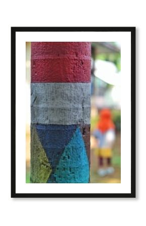 Close-up of a painted tree trunk with bold, colorful geometric designs. The blurred background adds depth, featuring a figure in orange, creating a mix of abstract art and outdoor elements
