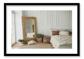 Bright and spacious bedroom with a white wall, an eco-wooden mirror and flower pots. Cozy bright rustic style bedroom, boho style bedroom.