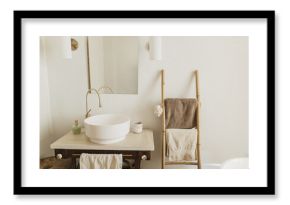 Stylish boho bathroom design. Modern ceramic sink with golden faucet on rustic stand with soap and ladder with towel on background of wall with mirror, modern bathroom interior.