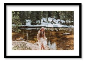 Side view of charming young long haired female in Nordic style boho clothes and wreath strolling along river shore in sunny early spring day with snowy forest landscape in background
