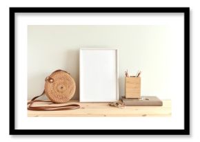 White blank frame mockup for poster or photo on wooden shelf, round rattan bag, boho style bracelets.