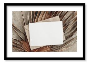 Summer stationery still life. Closeup of blank card mock-up and craft envelope on dry palm leaf. Grunge beige concrete background. Flat lay, top view. Tropical vacation concept. Moody boho design.