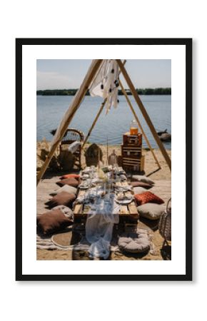 Set up wedding, serving table. Bachelorette party on beach. Boho style picnic. Decoration for celebration birthday. Lunch in romantic colors. Picnic with blanket, pillows, arch, garlands, wooden decor