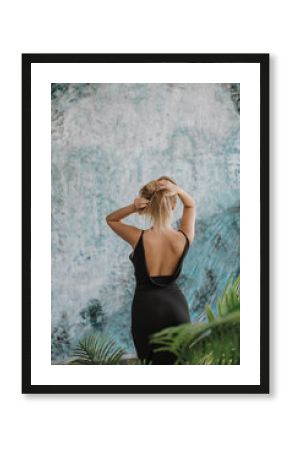 Back view of a blond woman in black backless dress, blu abstract wall in the background.