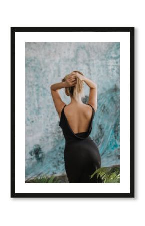 Back view of a blond woman in black backless dress, blu abstract wall in the background.