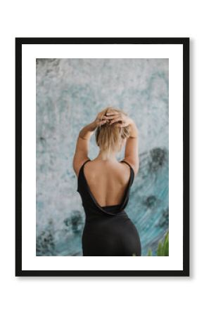 Back view of a blond woman in black backless dress, blu abstract wall in the background.