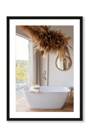 Comfortable bright bathroom with a boho-chic interior design, a free-standing white bath against the background of a panoramic window. hanging decor from dry grass. Soft selective focus.