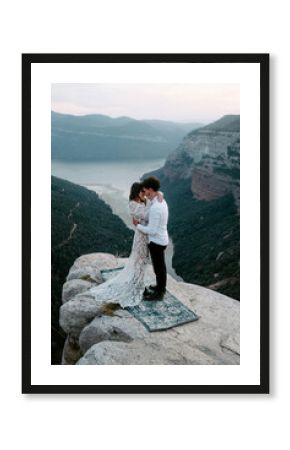 Side view of newlywed young man and woman in elegant clothes standing on top of rocky cliff of Morro de Labella embracing each other and kissing gently