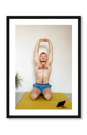 a man of European appearance is doing yoga online in a bright room at home. An athlete with a naked torso raises his arms up, performs a stretching exercise. 