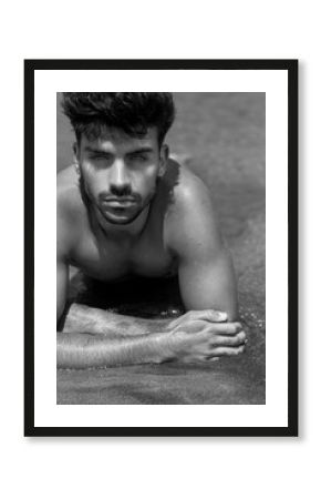 Retrato de chico hispano joven y guapo tirado en la arena. Fotografía en blanco y negro, primer plano. Hombre con barba y mirada seductora