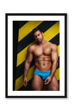 Naked muscular man in underwear at background with yellow and black lines. Male fitness model in studio. Brutal big guy posing in blue briefs.