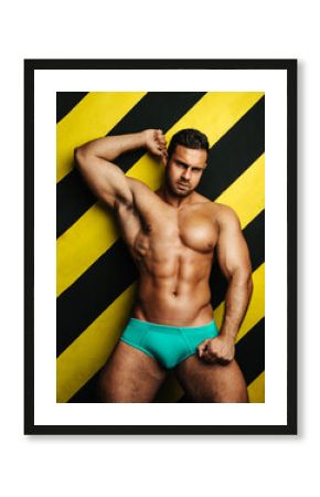 Naked muscular man in underwear at background with yellow and black lines. Male fitness model in studio. Brutal big guy posing in blue briefs.