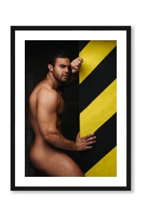 Naked man near wall. Male fitness model posing without wear in studio. Handsome guy showing his muscular body on black background.