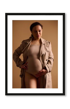 Attractive confident pregnant woman in lingerie and trench coat, stroking her belly, isolated on beige studio background