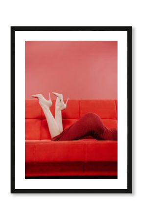 A girl with a beautiful shoe lays on a red sofa.Stylish girl in red dress is on a red sofa. Beautiful legs. Figure. Sexuality.Sexy woman's legs in high heels.Fashion, shoes concept.