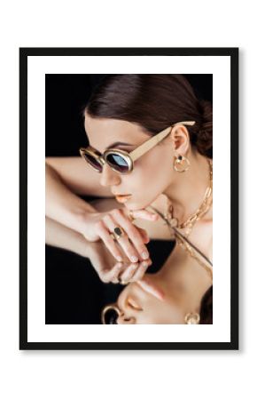 young naked woman in sunglasses, golden accessories lying on mirror isolated on black