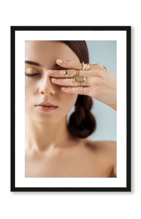 young naked woman with shiny makeup and closed eyes in golden rings hiding eye behind hand isolated on grey