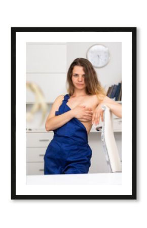 Portrait of naked woman cleaner in overall standing in office, covering her breast and looking at camera.
