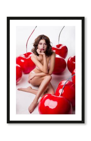 Elegant young fashion model in white lingerie sits in large cherries. Studio shot. Slim figure