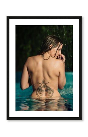 Naked woman with tattoos on her back in a swimming pool with wet skin and a waterfall and vegetation in the background.