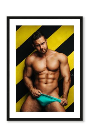 Naked muscular man in underwear at background with yellow and black lines. Male fitness model in studio. Brutal big guy posing in blue briefs.