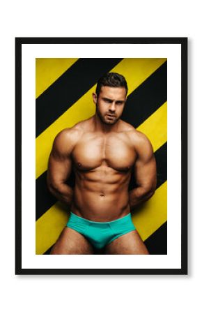 Naked muscular man in underwear at background with yellow and black lines. Male fitness model in studio. Brutal big guy posing in blue briefs.