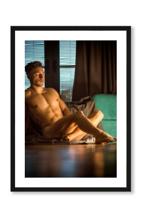 Handsome totally naked muscular young man sitting down on hardwood floor at home in seductive attitude, looking at camera