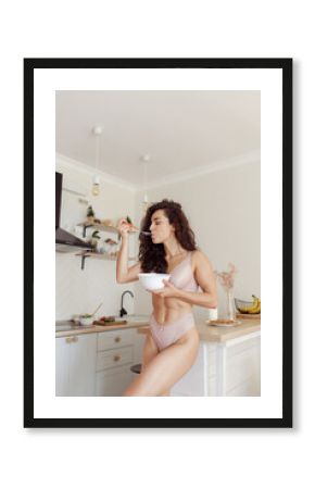 Vertical shot of sexy Caucasian pretty female standing in kitchen eating breakfast wearing underwear. Beautiful naked woman in lingerie eat cereals at home. Lazy morning concept