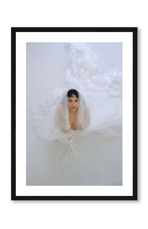 sexy young nude woman wrapped in translucent fabric, plastic foil, gazing upwards with a gentle, serene expression, creating an ethereal and peaceful atmosphere around her
