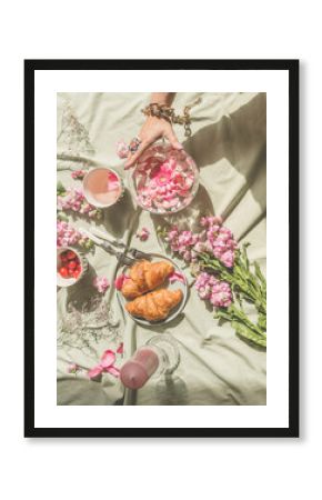 Aesthetic picnic at sun day with croissants, tea, bouquet of pink flowers and candles. Women hand holding glass vase with floating petals. Top view
