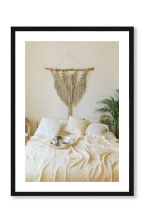 Modern romantic scandi boho style bedroom interior with decorative pillows, green plant and diy macrame wall panel. Light warm cozy comfortable home. jomo, weekend breakfast in bed concept.