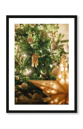 Stylish Christmas tree with modern baubles, boho ornaments and golden lights and glowing star. Atmospheric festive scandinavian room in Christmas night. Magic time. Merry Christmas!