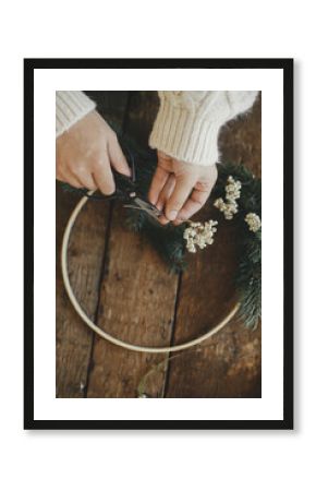 Hands in cozy sweater cutting herb with scissors for modern boho wreath with fir branches and wooden hoop on rustic table. Atmospheric moody image. Making stylish christmas wreath