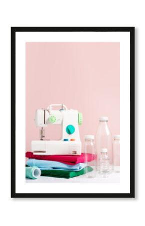 Plastic bottles, sewing machine and folded t-shirts still life on pink, concept of clothes made from recycled plastic
