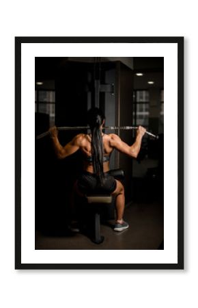 Front view of strong muscular back of sports female bodybuilder who trains on the simulator.