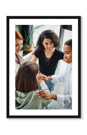 high angle view of diverse group of pleased multicultural women joining hands near positive motivation coach during session in consulting room, moral support and mental wellness concept