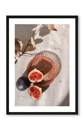 Aesthetic minimal autumn food still life. Whole and cut fig fruits on neutral beige linen tablecloth background with plate, dried leaves and harsh sunlight shadows