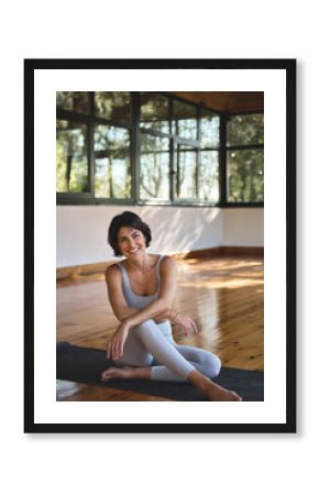 Young happy fit sporty slim Hispanic woman yoga coach, stretching sport trainer wearing grey top and pants sitting on mat in modern yoga gym studio in nature park looking at camera. Vertical portrait.