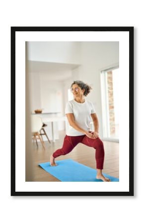 Sporty fit middle aged woman standing on mat at home doing fitness exercises morning routine. Happy healthy mature 50 years old lady enjoying casual workout or yoga in living room, vertical photo.