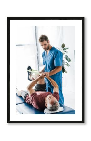 handsome doctor holding leg of mature patient exercising with elastics