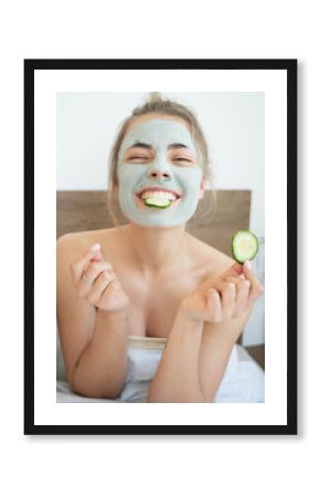 Happy beautiful young woman with cosmetic clay mask on her face eating cucumber and laughing in her bedroom at home. Self care, stay at home. Face skin care concept.