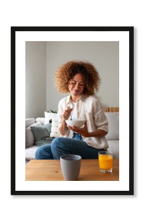 Vertical portrait of young multiracial woman eating breakfast sitting on sofa at home living room.