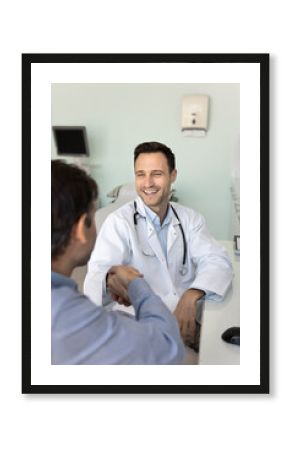 Positive satisfied male doctor giving handshake to healthy patient, sharing good news after getting medical examination results, shaking hands with man at work table, smiling, laughing