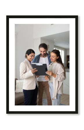 Happy young Indian insurance agent showing and explaining agreement to couple of clients at home. Realtor and customer reviewing paper document on pad for signing. Vertical shot