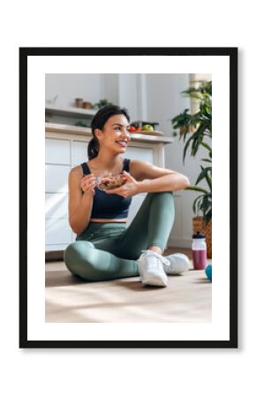 Athletic woman eating a healthy bowl of muesli with fruit sitting on floor in the kitchen at home