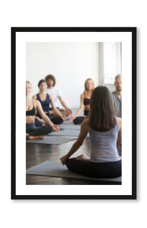 Group of young sporty people practicing yoga lesson with instructor, sitting in Padmasana exercise, Lotus pose, working out, indoor full length, studio, students training in club, rear view at teacher