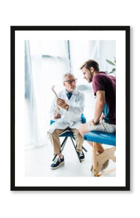 bearded doctor in glasses holding spine model and looking at patient